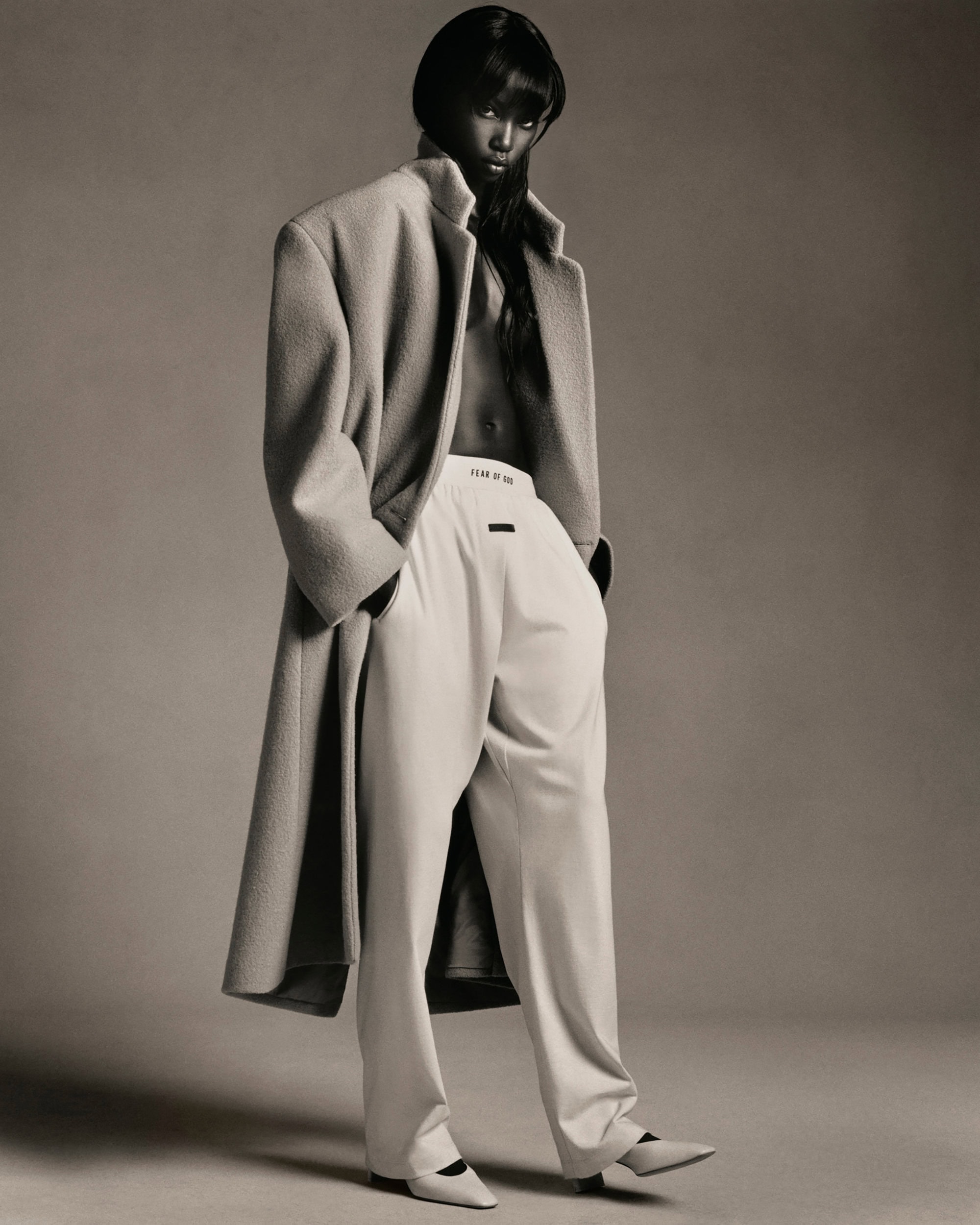 Elegant fashion shot of an adult woman in an oversized wool coat and white pants, standing in a neutral studio setting, minimalist style.