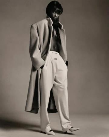 Elegant fashion shot of an adult woman in an oversized wool coat and white pants, standing in a neutral studio setting, minimalist style.