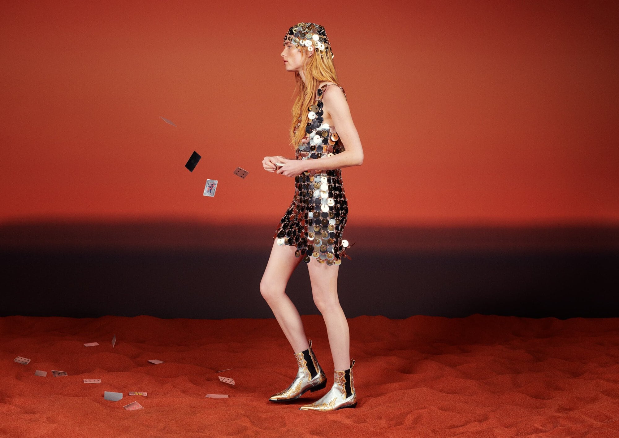 A fair-skinned adult female model stands in a shiny dress and boots, with playing cards suspended in the air on a gradient orange background.