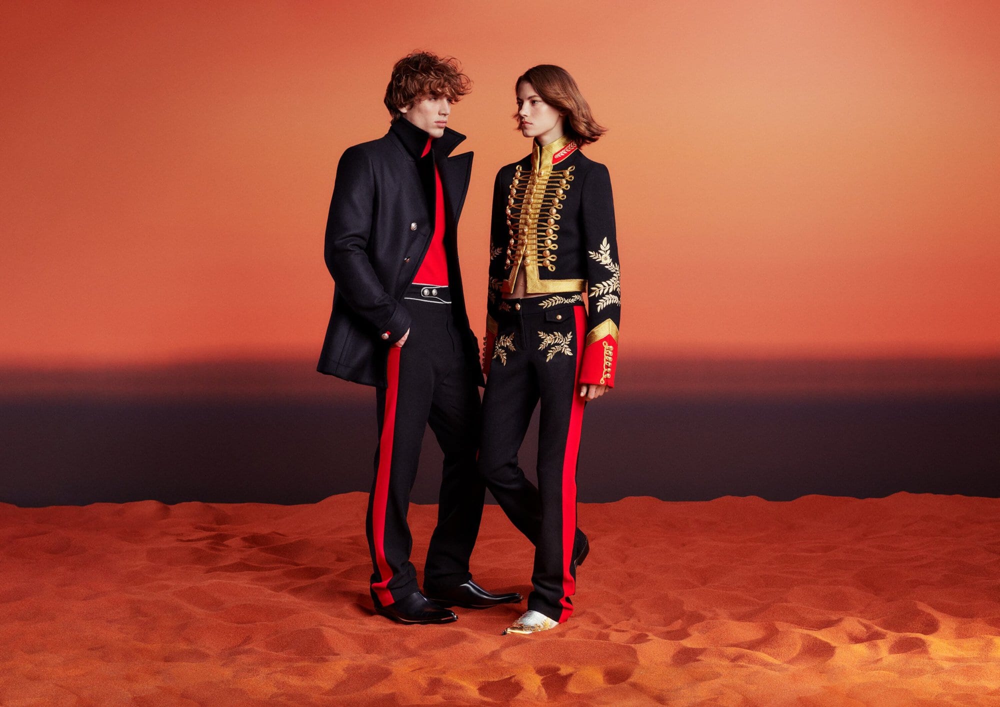 Two models pose in black and red military-style outfits on a textured orange surface with a blurred gradient background.