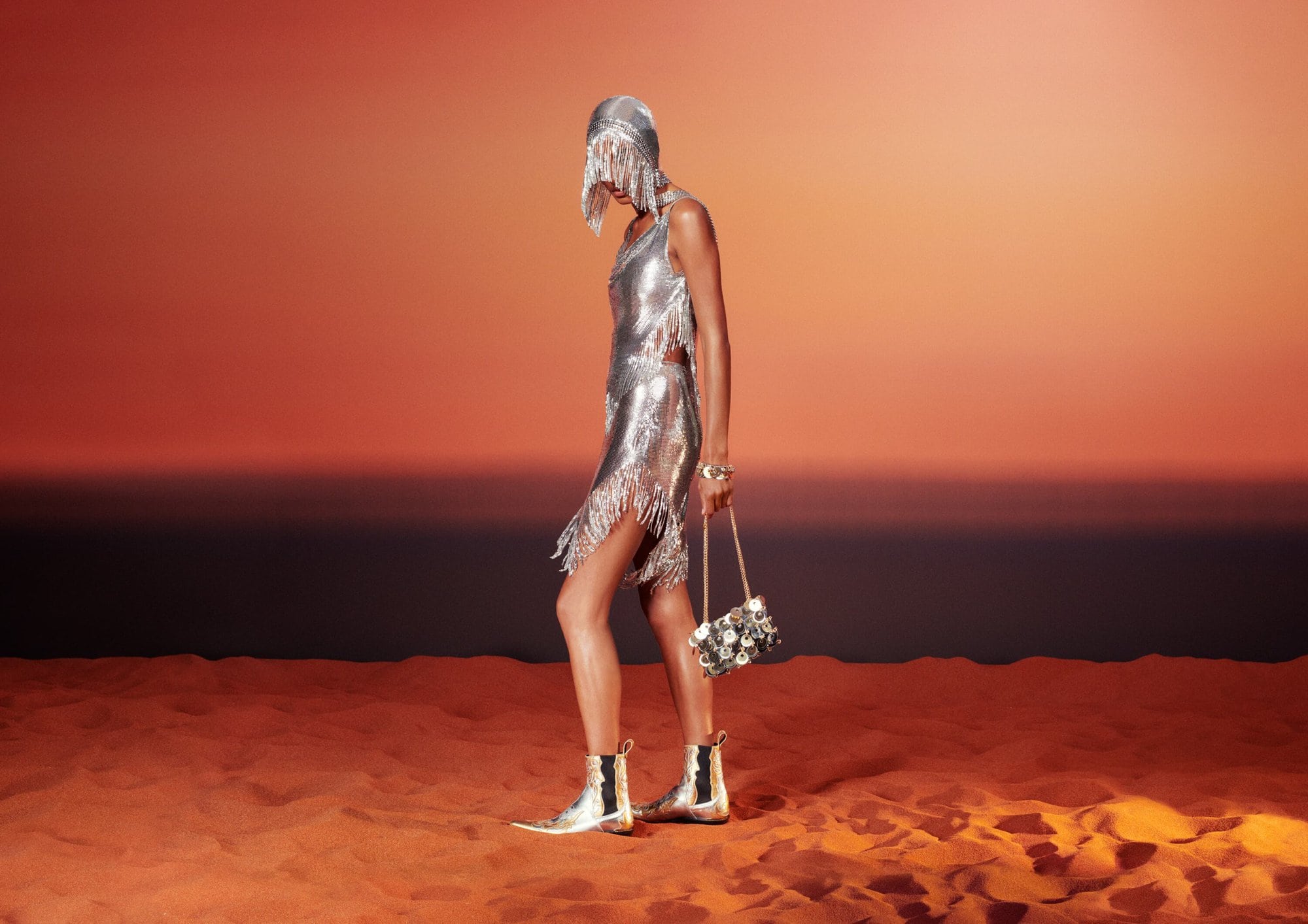 A woman in a silver, fringed dress and matching headdress stands in a desert, holding a silver bag and wearing silver boots.