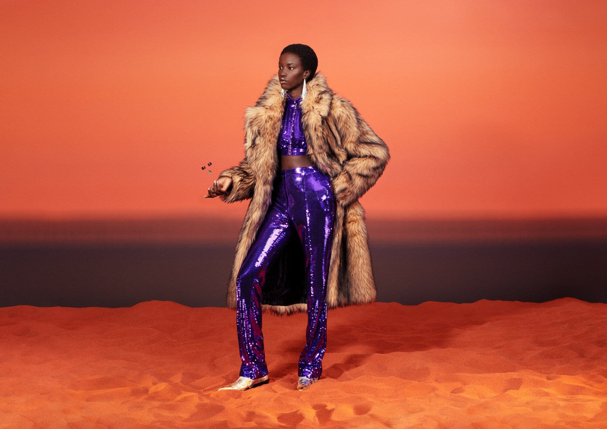 An adult female model poses in a furry coat and purple sequin outfit against a gradient orange backdrop.  The model is standing on a textured surface.