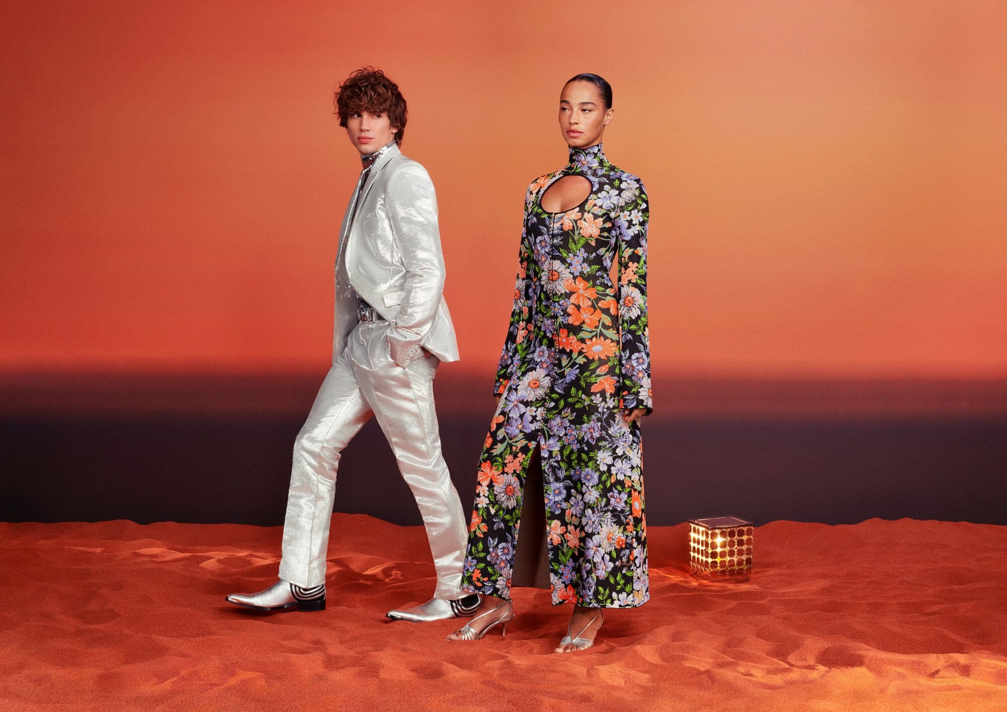 Two fashion models showcase clothing in an indoor studio setting. One wears a silver suit; the other, a floral dress, against an orange backdrop.