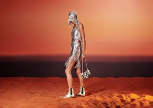 Model wearing a metallic silver chainmail fringe dress, headdress, and boots on orange sand against a gradient sunset backdrop.
