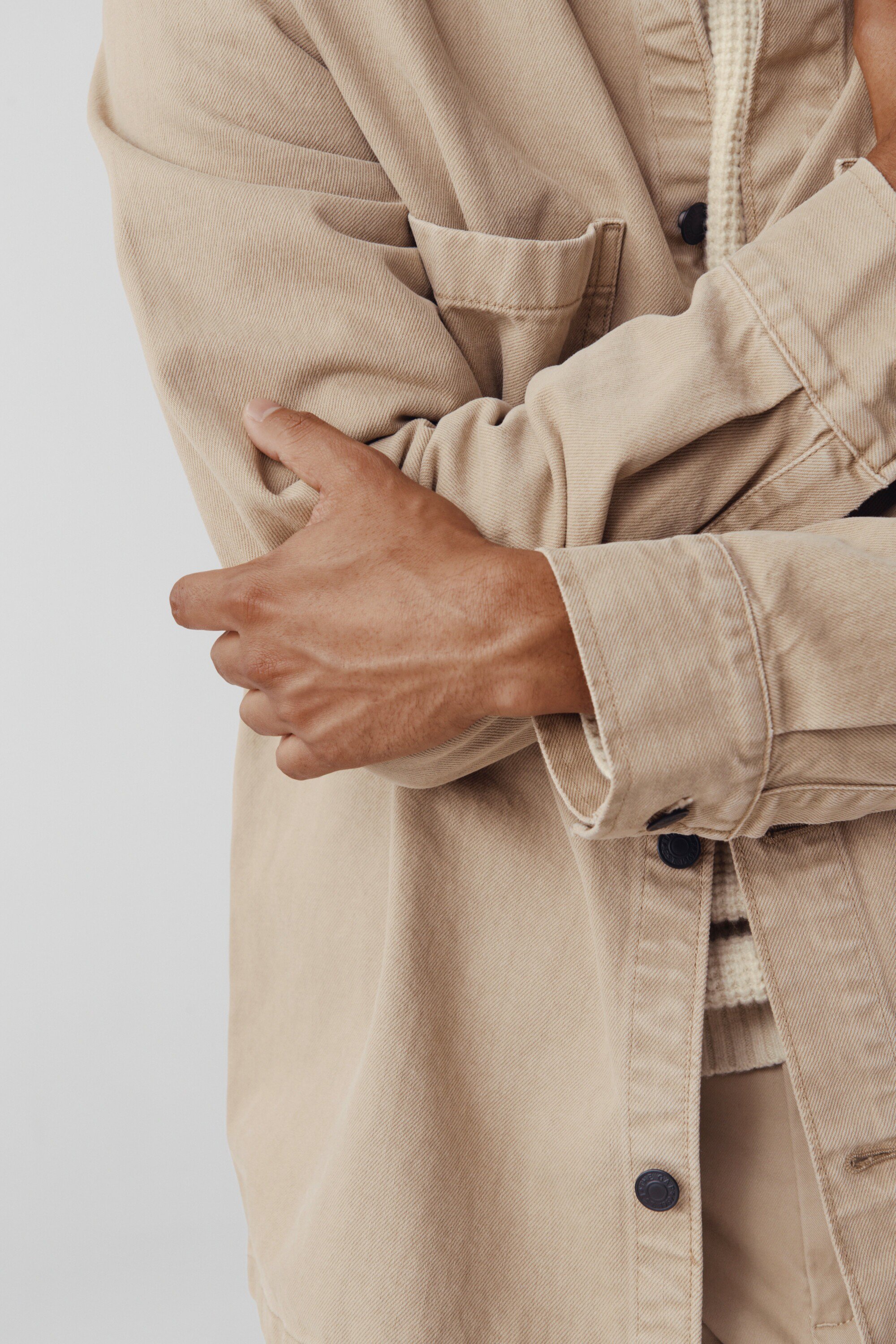 Close-up studio shot of adult male wearing beige denim jacket and striped top with arms crossed against a white background.