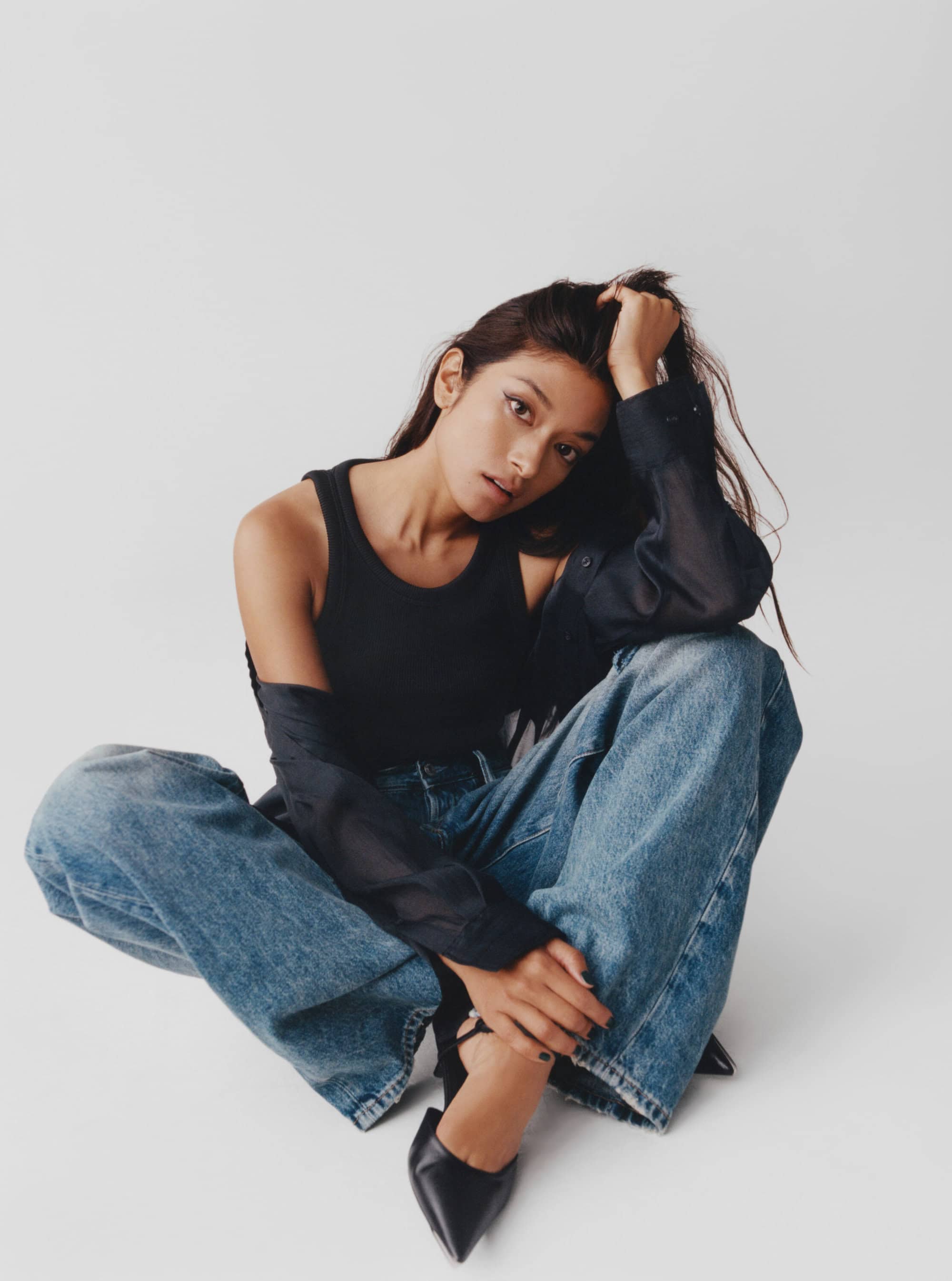 A woman poses in a black top and blue jeans, seated on a white floor, one hand on her head, and wearing black high heel shoes.
