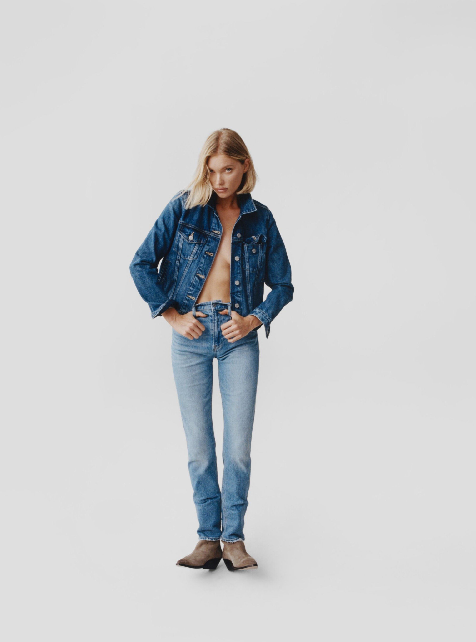 Blonde woman in denim jacket and jeans stands in a white photo studio, hands in pockets, looking down, with taupe suede ankle boots.