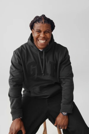 Jon Gray smiling while wearing a dark Gap hoodie and black trousers against a white seamless background.