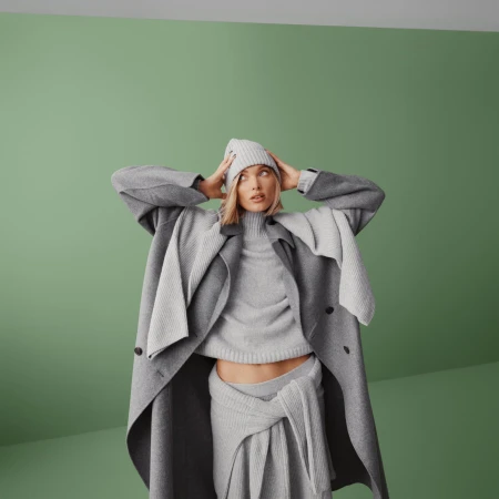 Elsa Hosk wearing a monochromatic grey knit sweater, beanie, and long coat against a muted green studio wall.