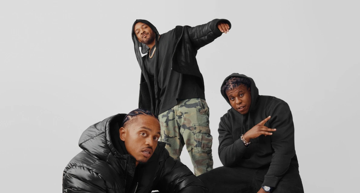 Three stylish adult males pose indoors against a neutral backdrop. One is standing wearing camo pants, while two are seated in dark jackets.