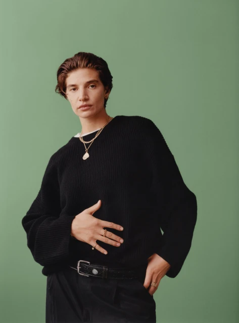 A woman with short brown hair poses indoors wearing a black sweater, black pants, rings, and necklaces against a green backdrop.