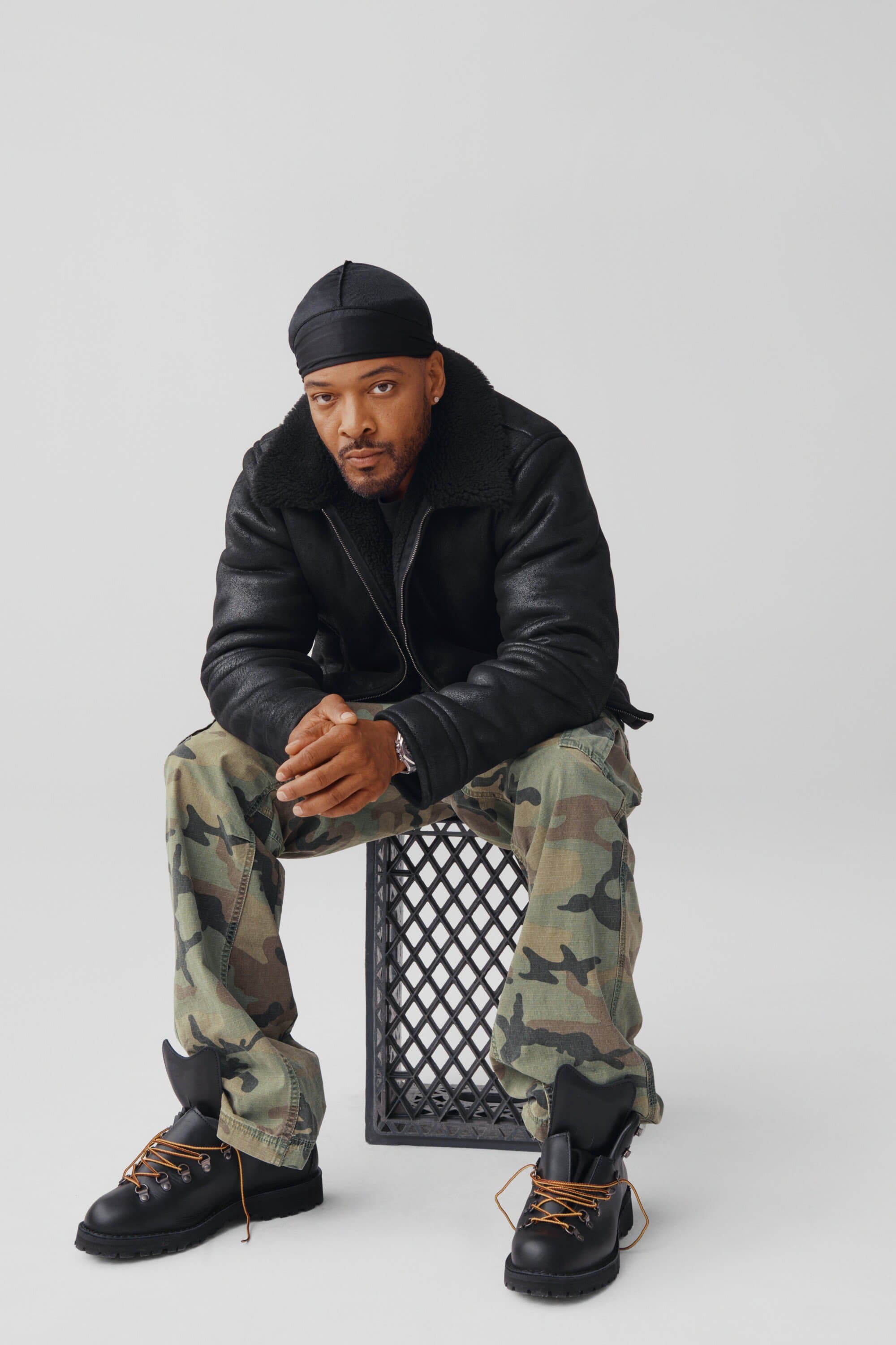 Adult male seated on a black plastic crate. He wears a black shearling jacket, camouflage pants, black boots and a black durag.
