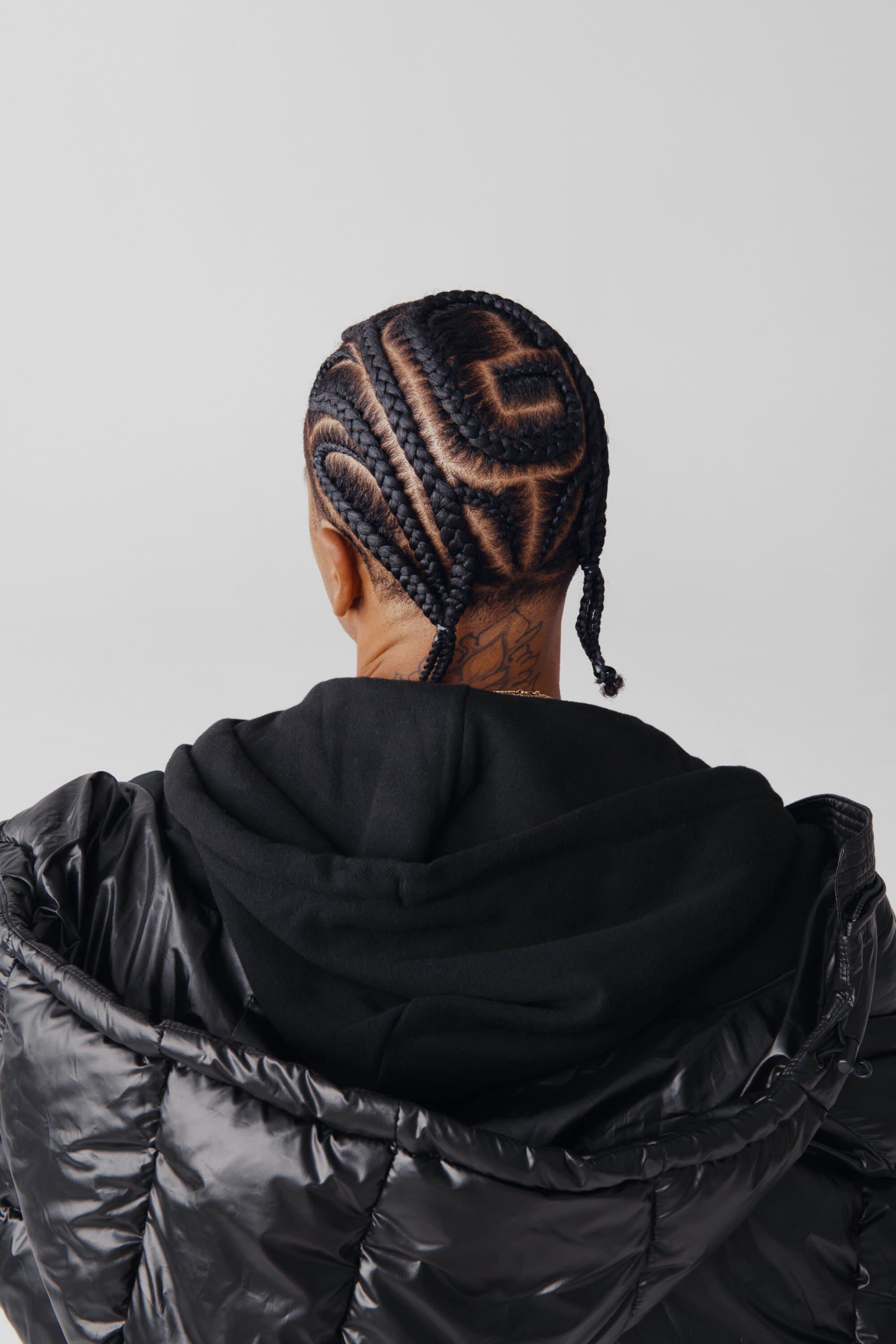 Back view of a person with intricate cornrow braids and a black puffer jacket against a plain white backdrop in a studio setting.