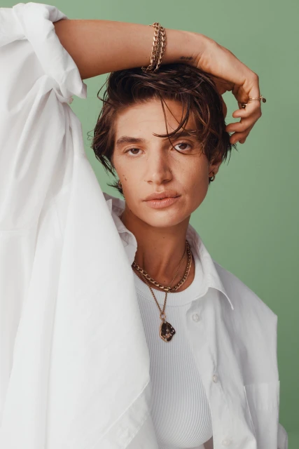 Portrait of a fashionable short-haired woman in a white shirt with gold jewelry, posing in front of a light green background.