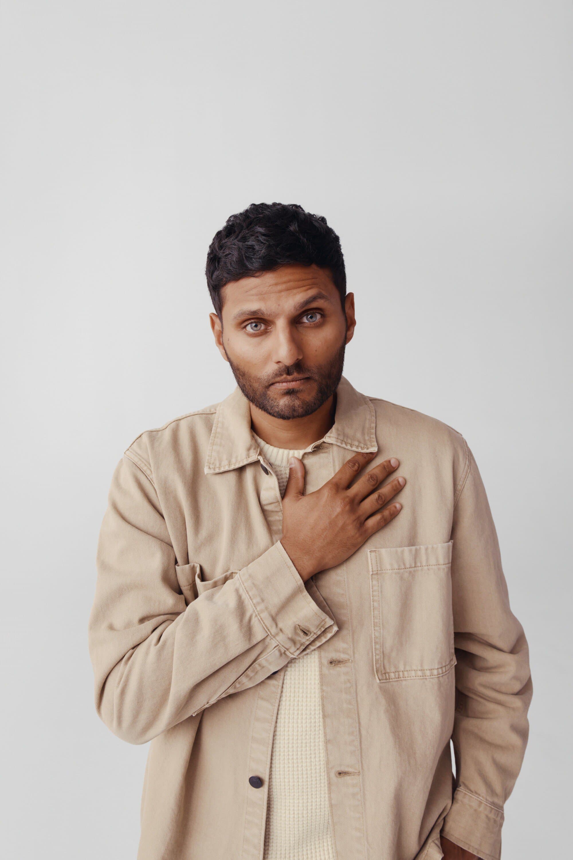 Portrait of a brunette adult male with hand on chest, wearing a beige jacket against a neutral gray backdrop in a studio setting.