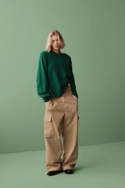 A woman with short blonde hair poses in a green sweater and baggy beige cargo pants against a solid green backdrop in a studio shot.