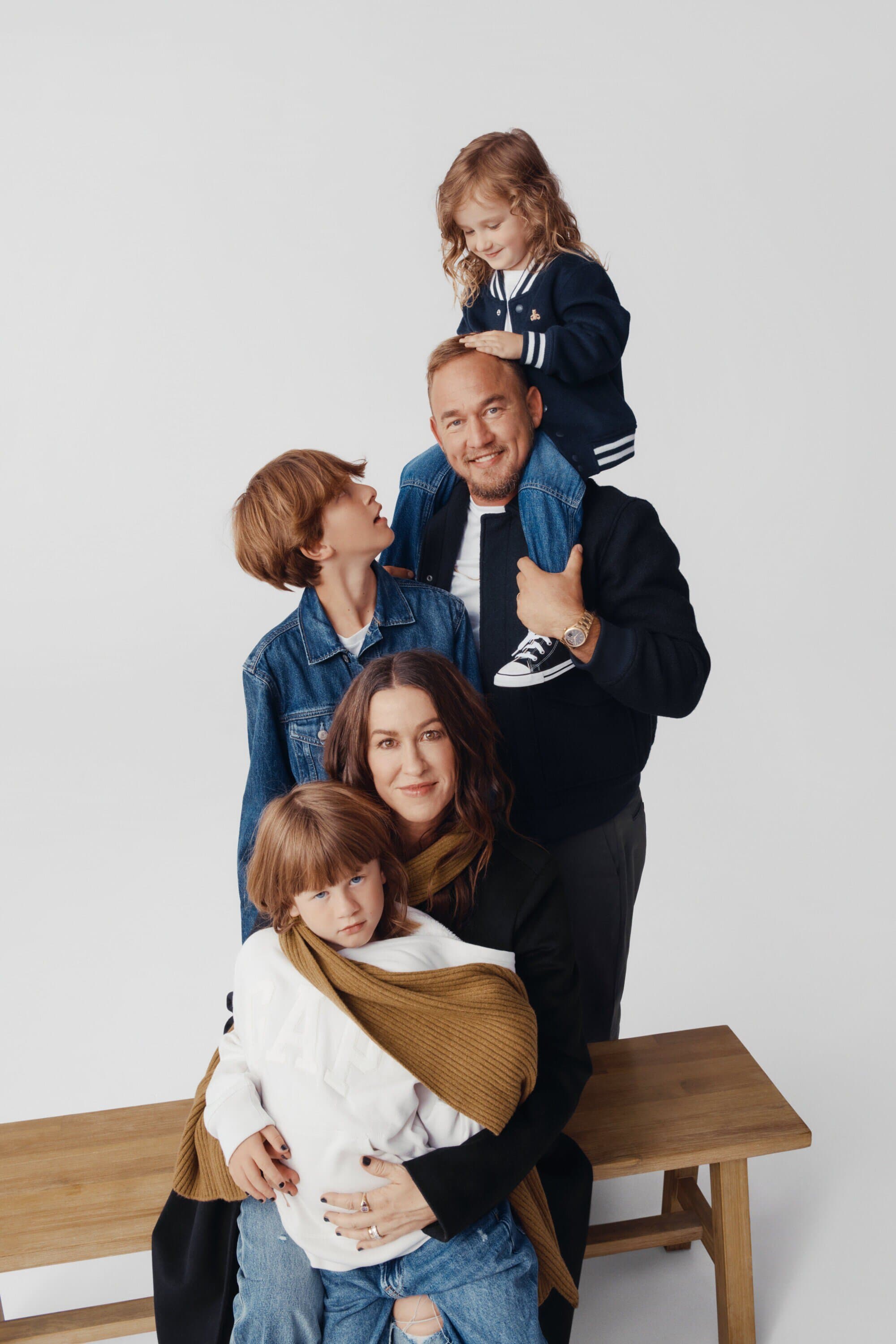 Family portrait of mother, father, daughter on shoulders, two sons, seated on wooden bench in front of a plain white background in studio lighting.