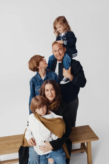 Family portrait of mother, father, daughter on shoulders, two sons, seated on wooden bench in front of a plain white background in studio lighting.
