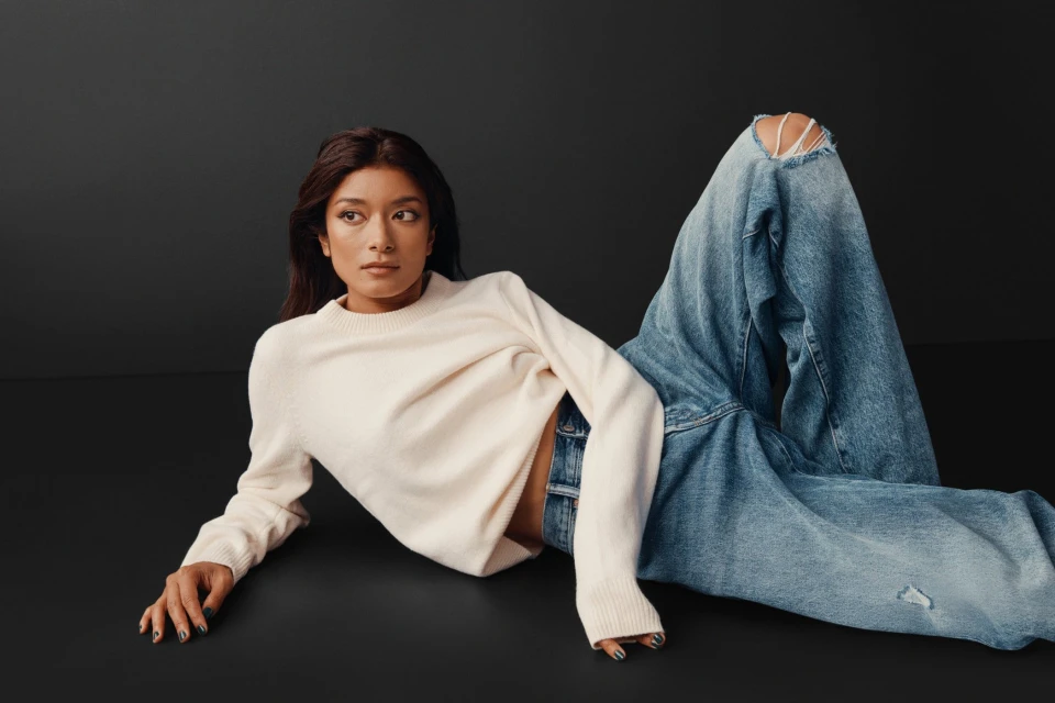 Woman in cream sweater and wide-leg ripped jeans poses on a dark grey seamless backdrop in a fashion editorial-style portrait.