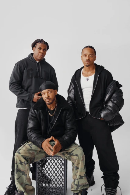 Three men pose in a photo studio. Two stand and one sits on a plastic crate, all wearing streetwear clothing, and looking at the camera.