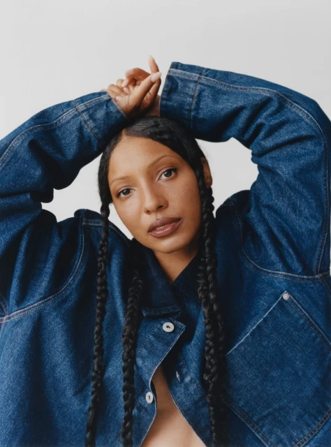 Close-up portrait of an African-American woman in a blue denim jacket with braided hair, posing against a white background in a photo studio.