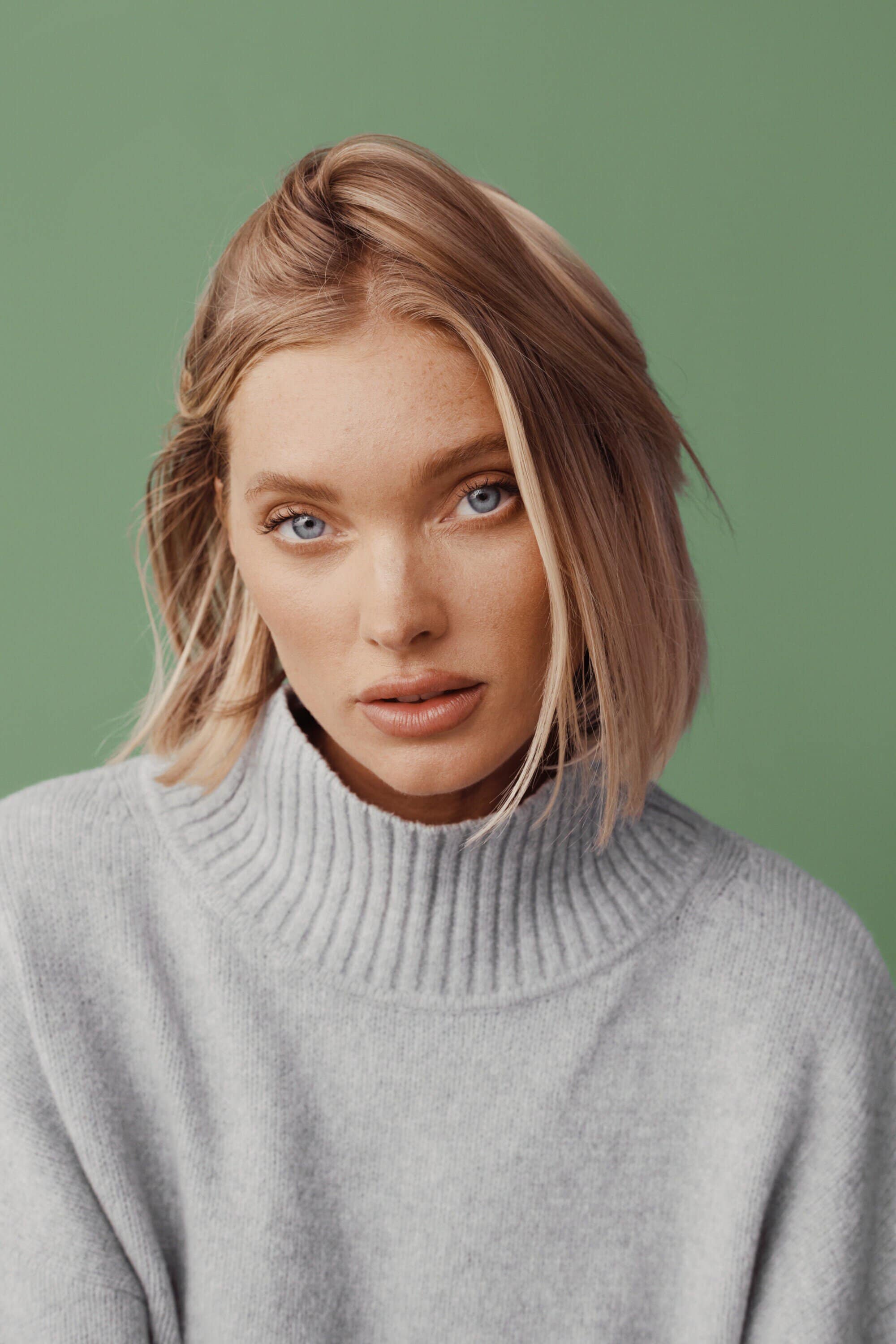 Close-up portrait of an adult woman with blonde hair wearing a grey turtleneck sweater against a light green background in a photo studio.