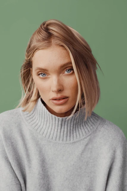 Close-up portrait of an adult woman with blonde hair wearing a grey turtleneck sweater against a light green background in a photo studio.