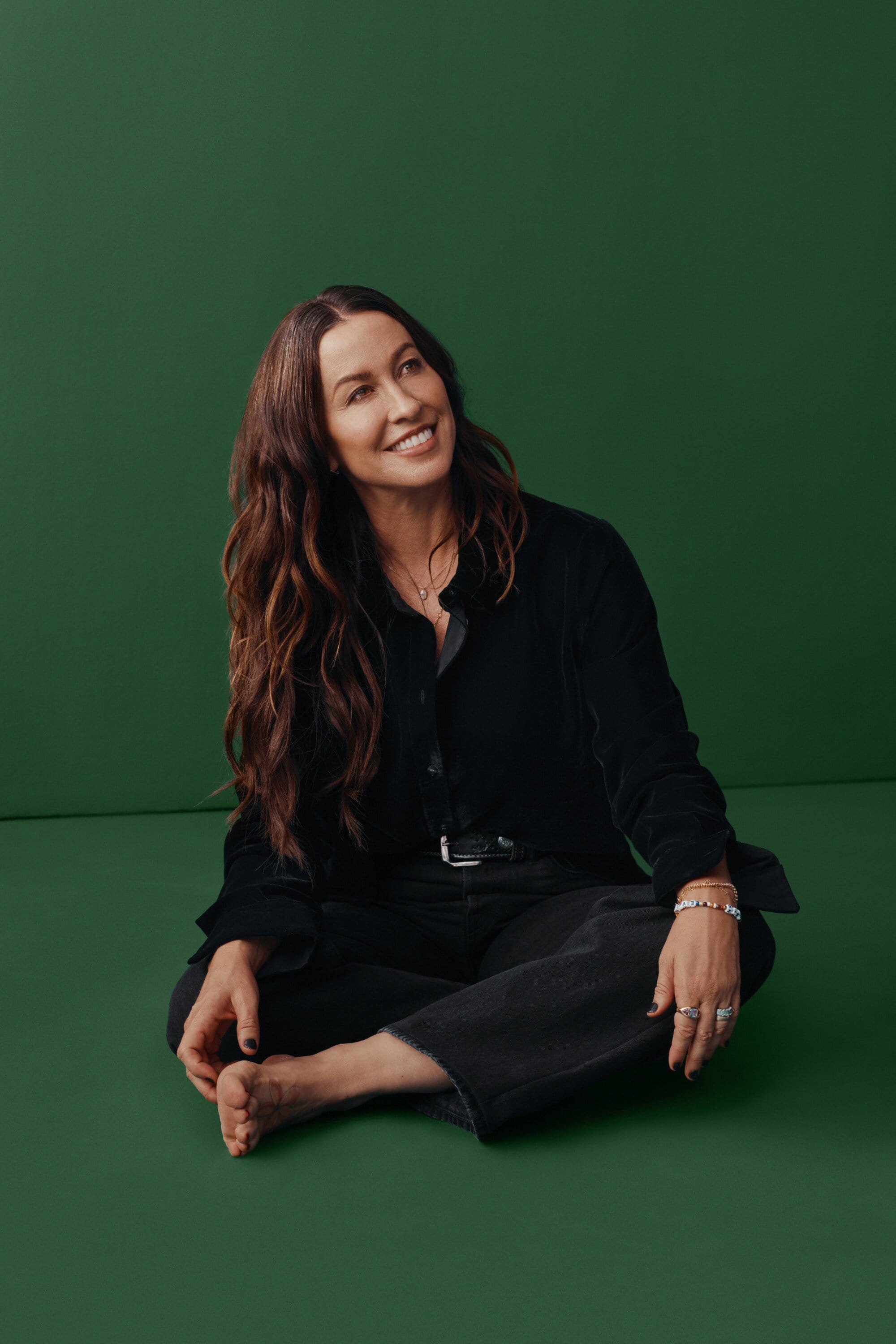 Attractive woman with long brunette hair sits cross-legged in front of a green backdrop, smiling and looking upward in a studio shot.