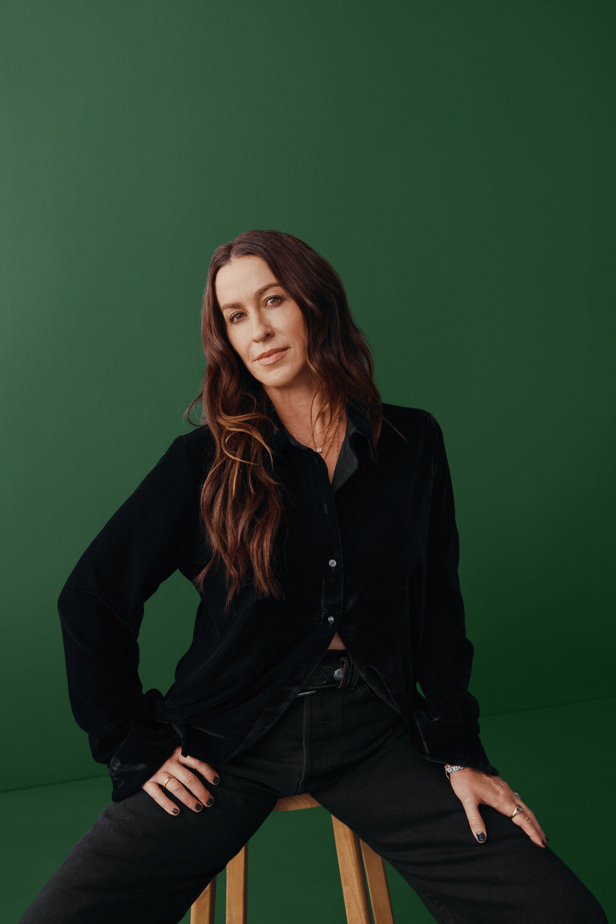 Portrait of woman with long brown hair, wearing a black velvet shirt and jeans, sitting on a stool against a green backdrop.