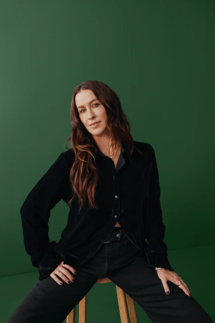 Portrait of woman with long brown hair, wearing a black velvet shirt and jeans, sitting on a stool against a green backdrop.