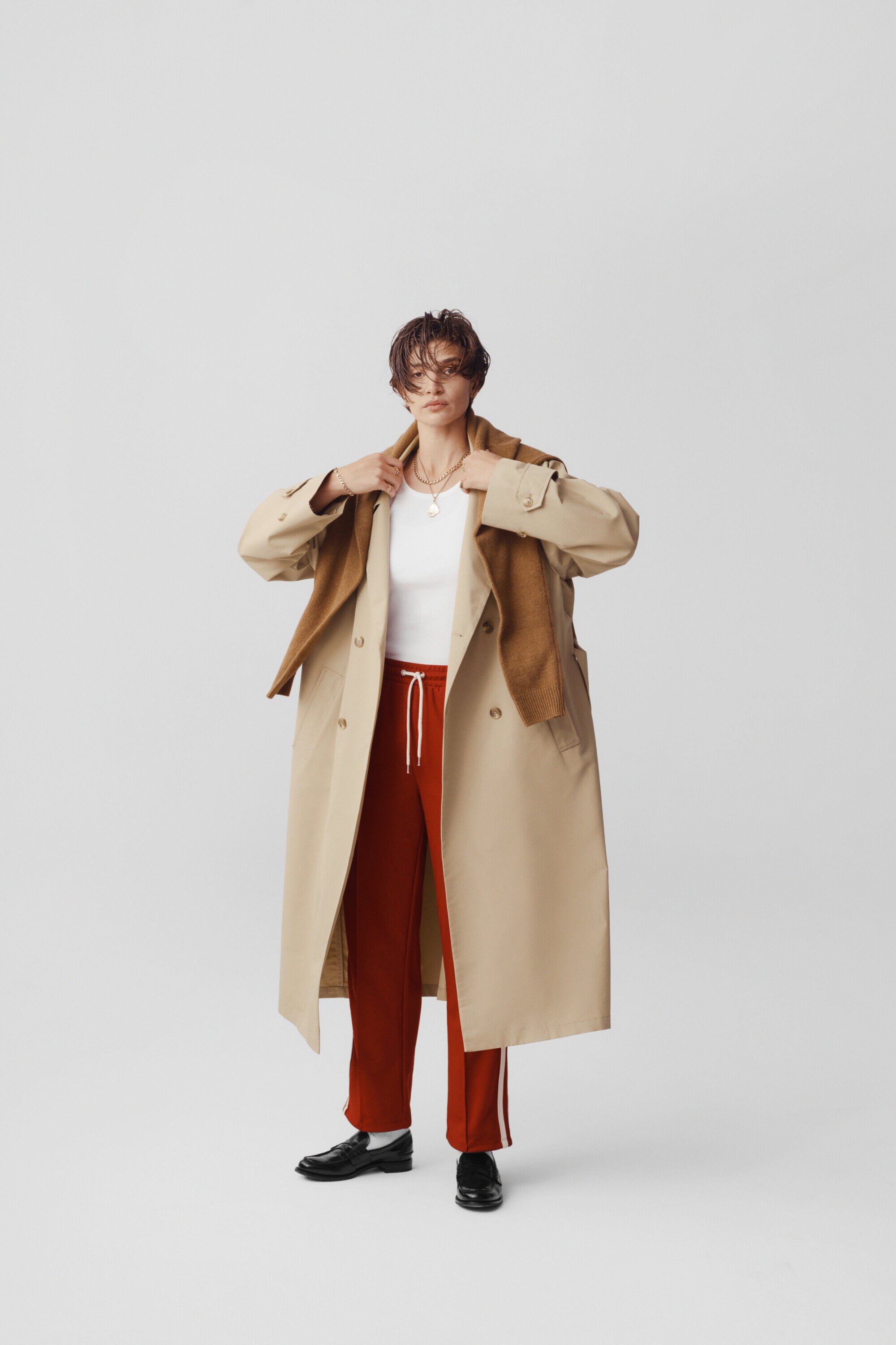 Full shot of a woman in a beige trench coat and red pants standing in a white photo studio, looking at the camera with a neutral expression.