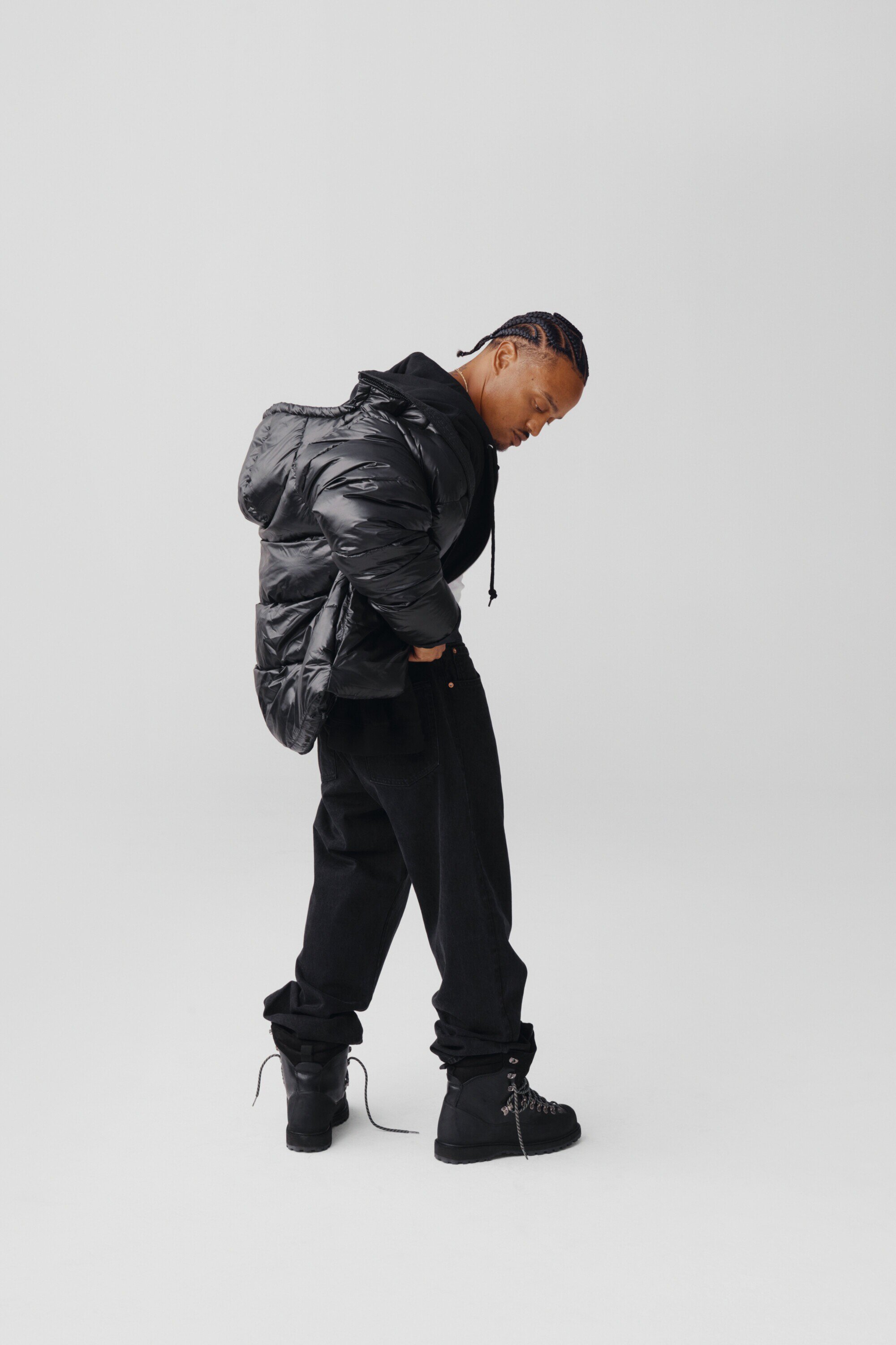 Man in a black puffer jacket and jeans poses in a studio with a clean white background and soft, neutral lighting, creating a sleek, modern look.