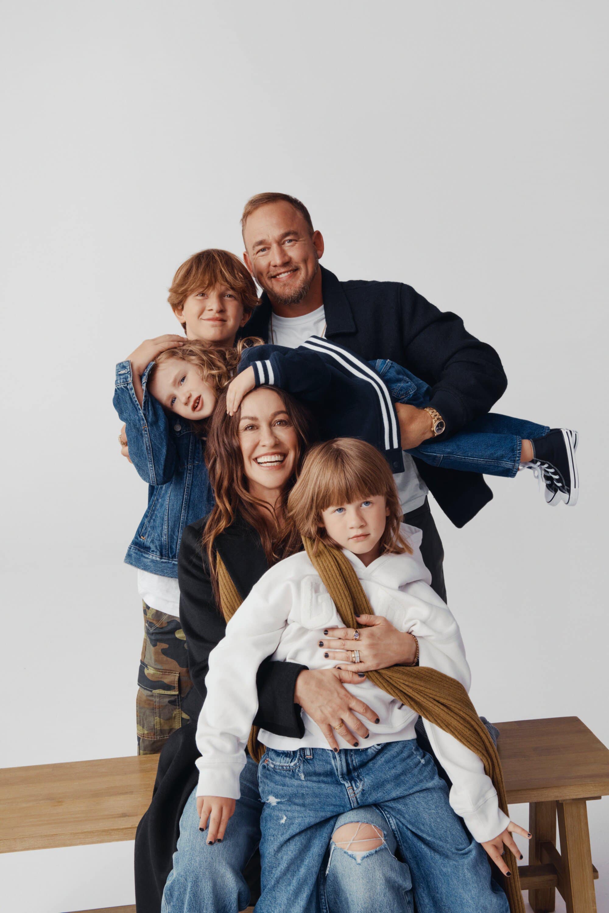 Family portrait featuring a mother, father, and three children in a photo studio with a neutral background, casually dressed, and smiling.