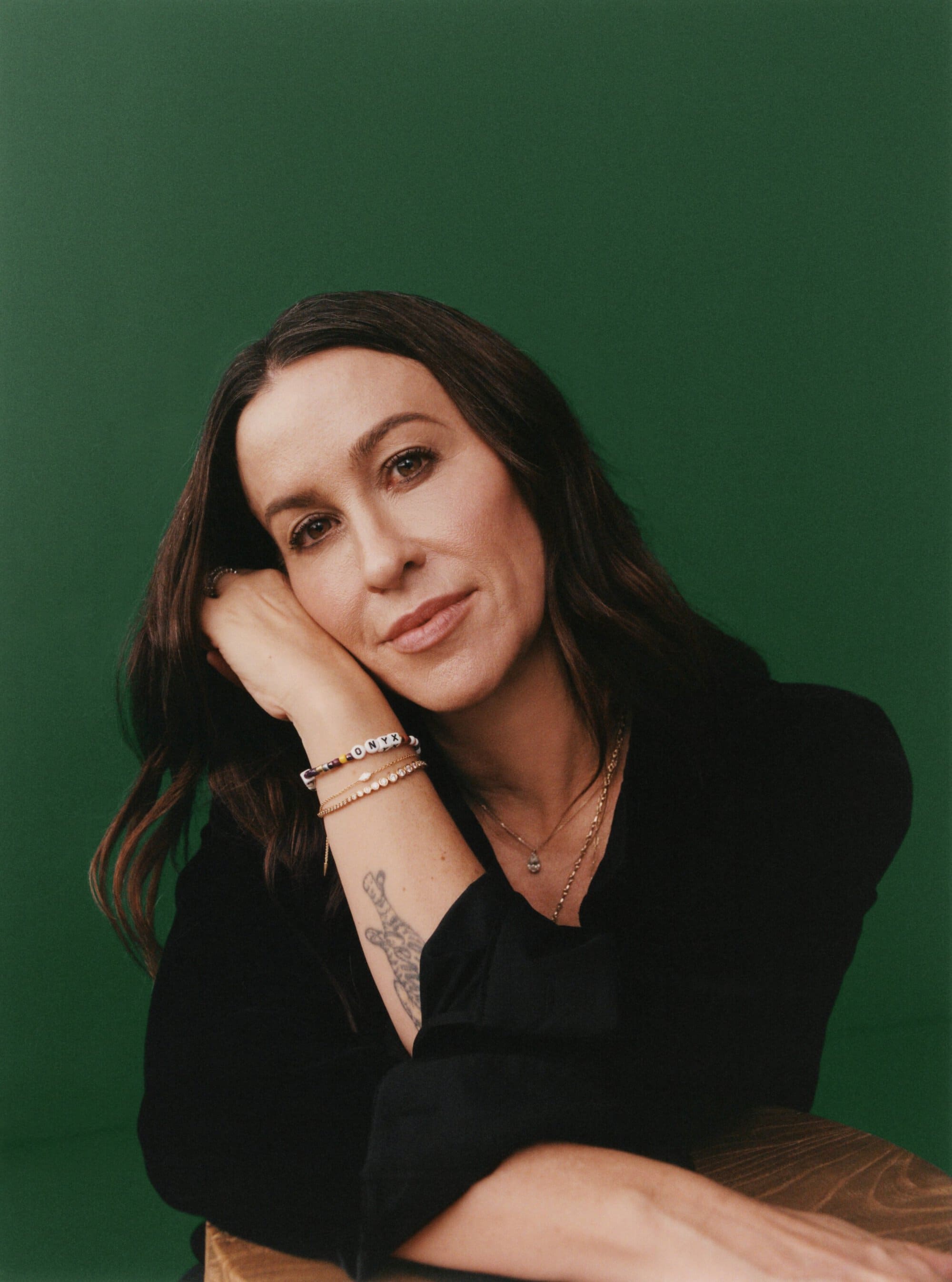 Portrait of an adult woman with dark hair in a black blouse, resting her head on her hand, adorned with bracelets against green background.
