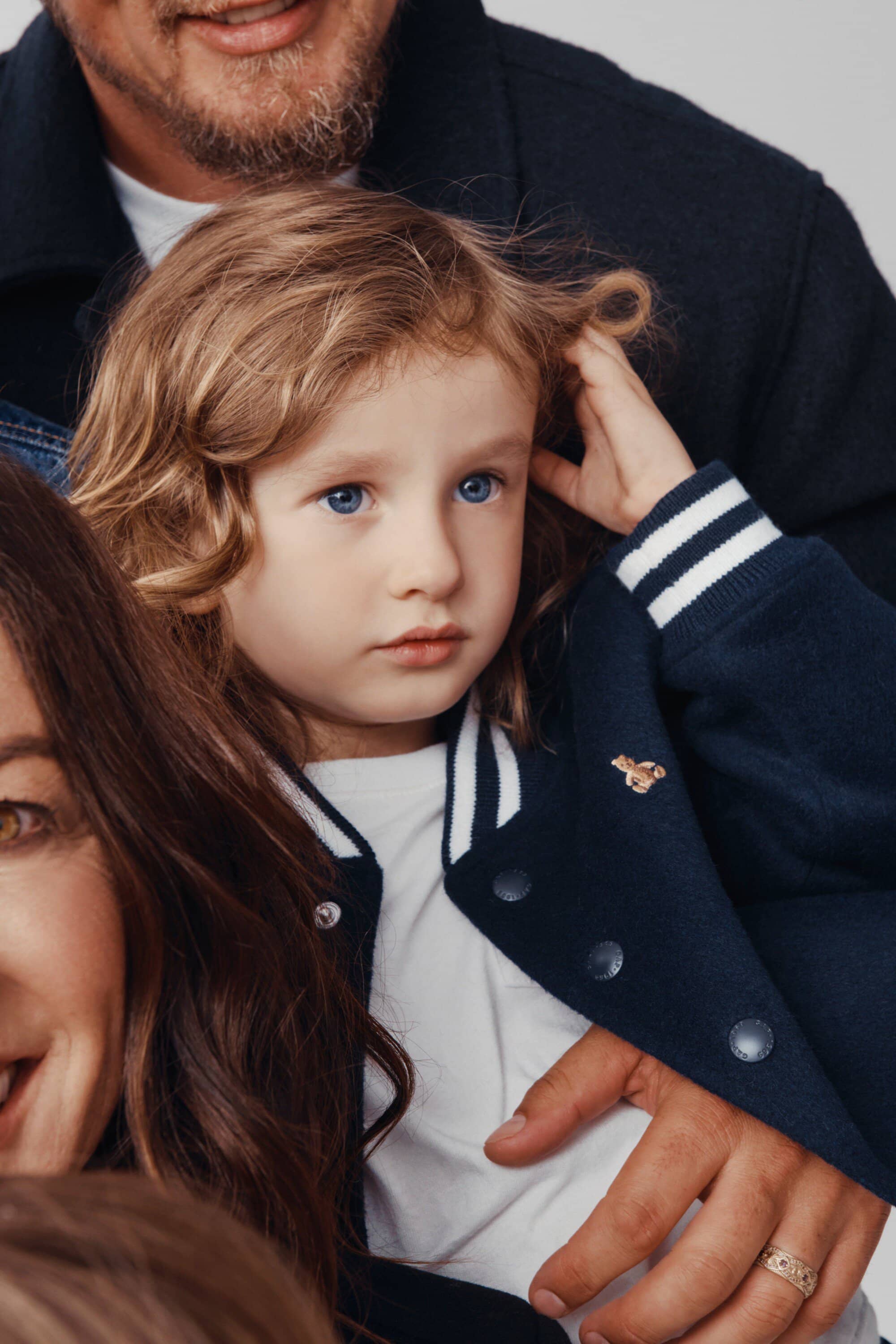 Close up of a family portrait featuring a young child with blonde curly hair in a jacket, with a blurred father and mother behind.