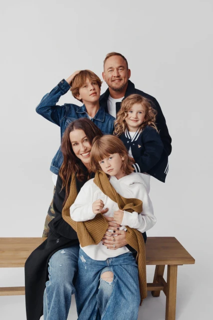 Family portrait in a photo studio. The group of five people are smiling and posing in casual outfits against a white backdrop.