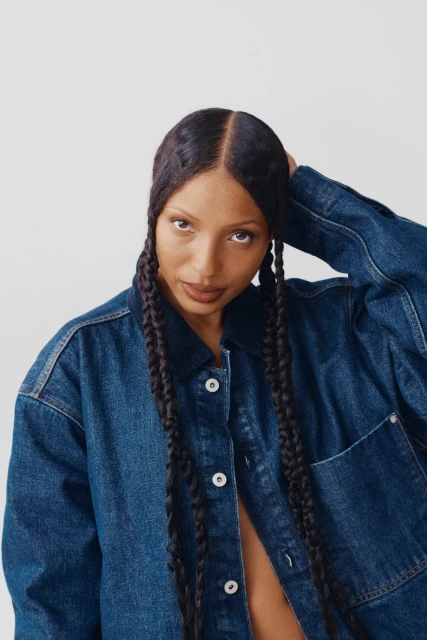 A woman with long braids poses in a denim jacket against a white background, looking directly at the camera in a close-up shot.
