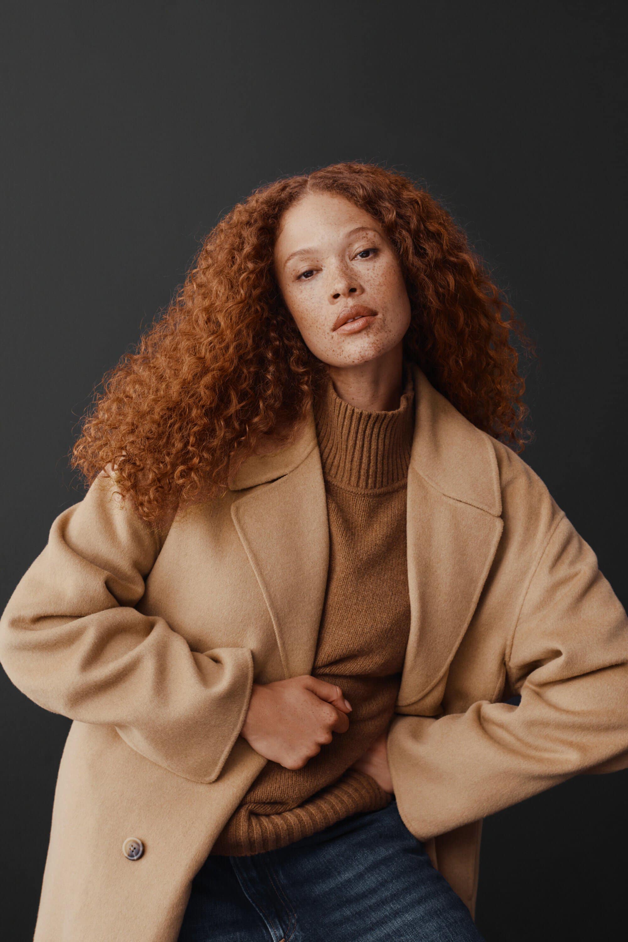A woman with red curly hair wears a tan coat and turtleneck with denim jeans in a studio portrait against a dark grey backdrop.