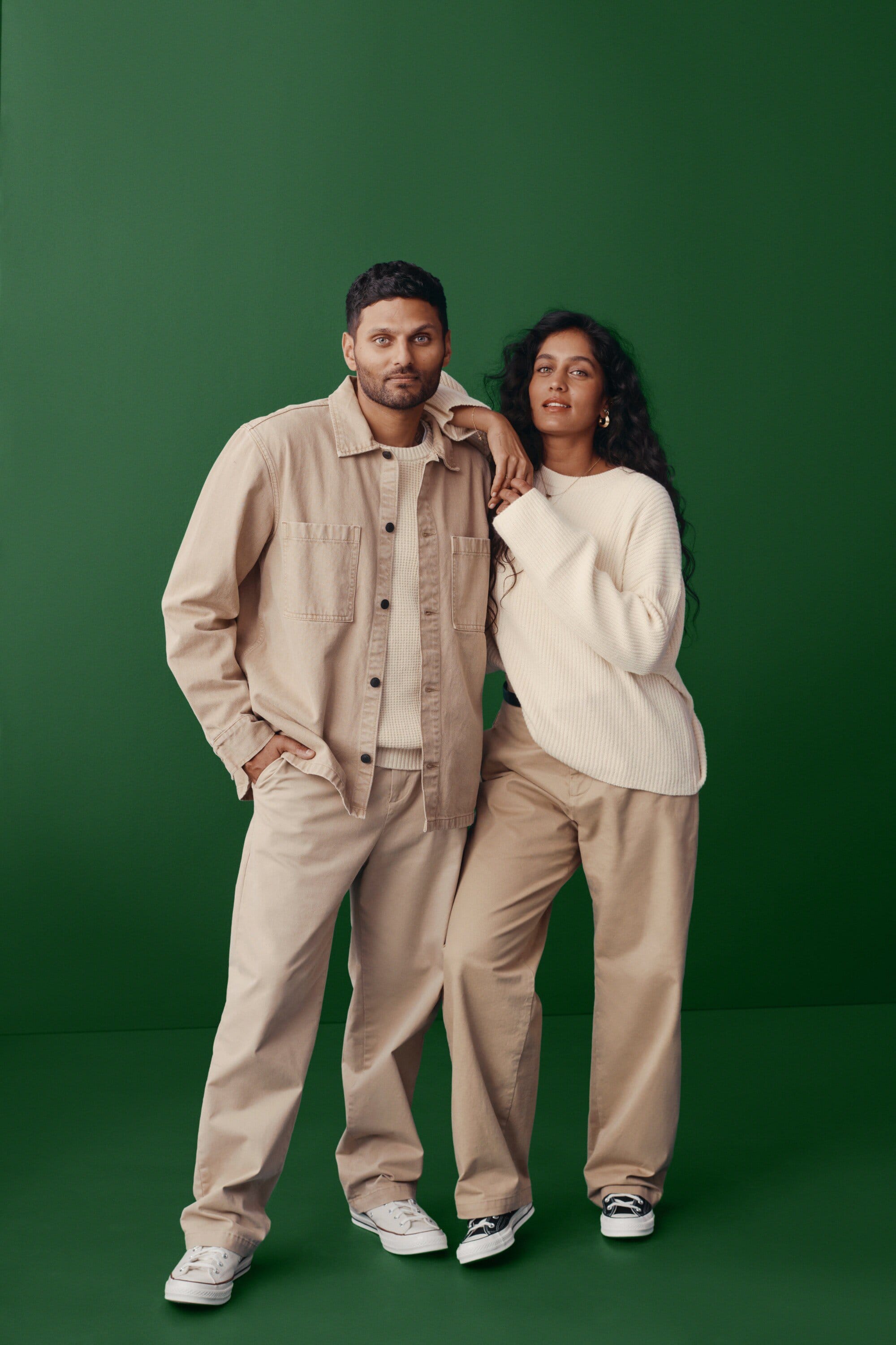 Two people pose against a green backdrop, the adult male in beige and the adult female in cream colored casual clothing.
