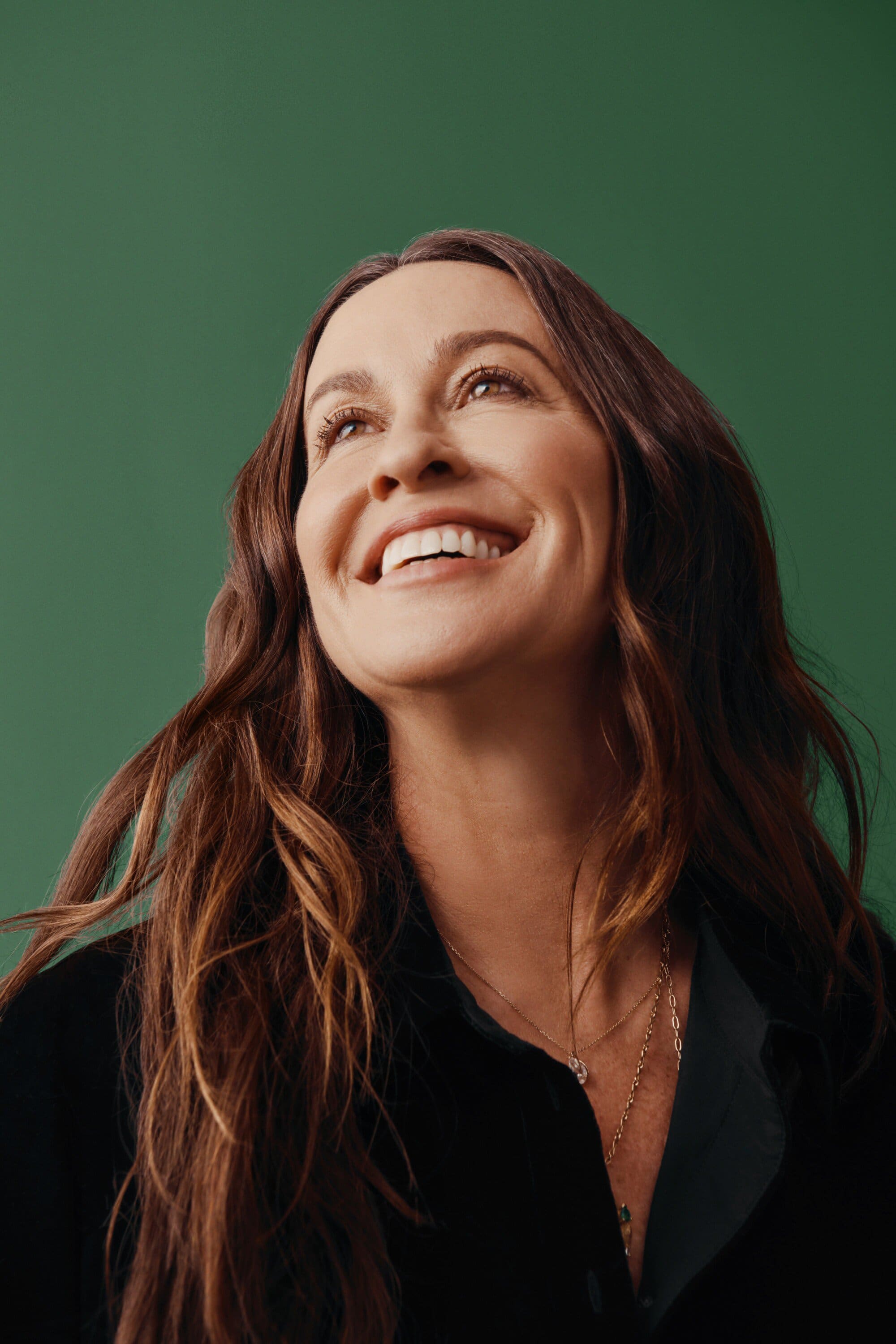 Close-up studio shot of a smiling adult woman with long wavy brunette hair, wearing a black top against a green background.