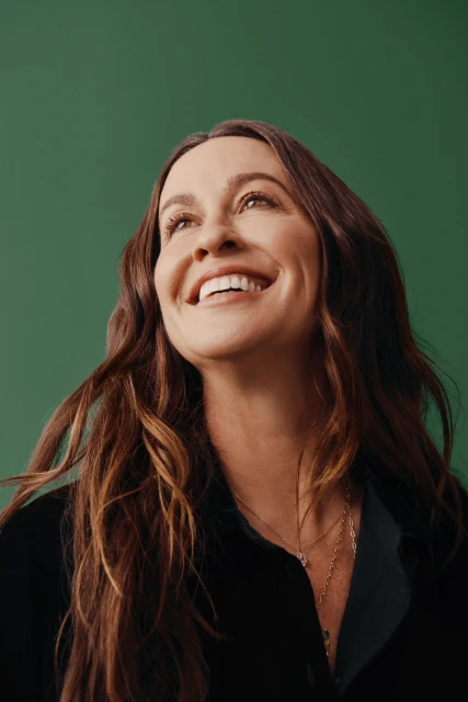 Close-up studio shot of a smiling adult woman with long wavy brunette hair, wearing a black top against a green background.
