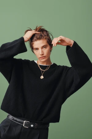 Model with short brown hair in an oversized black knit sweater and layered gold necklace posing against a muted green backdrop.