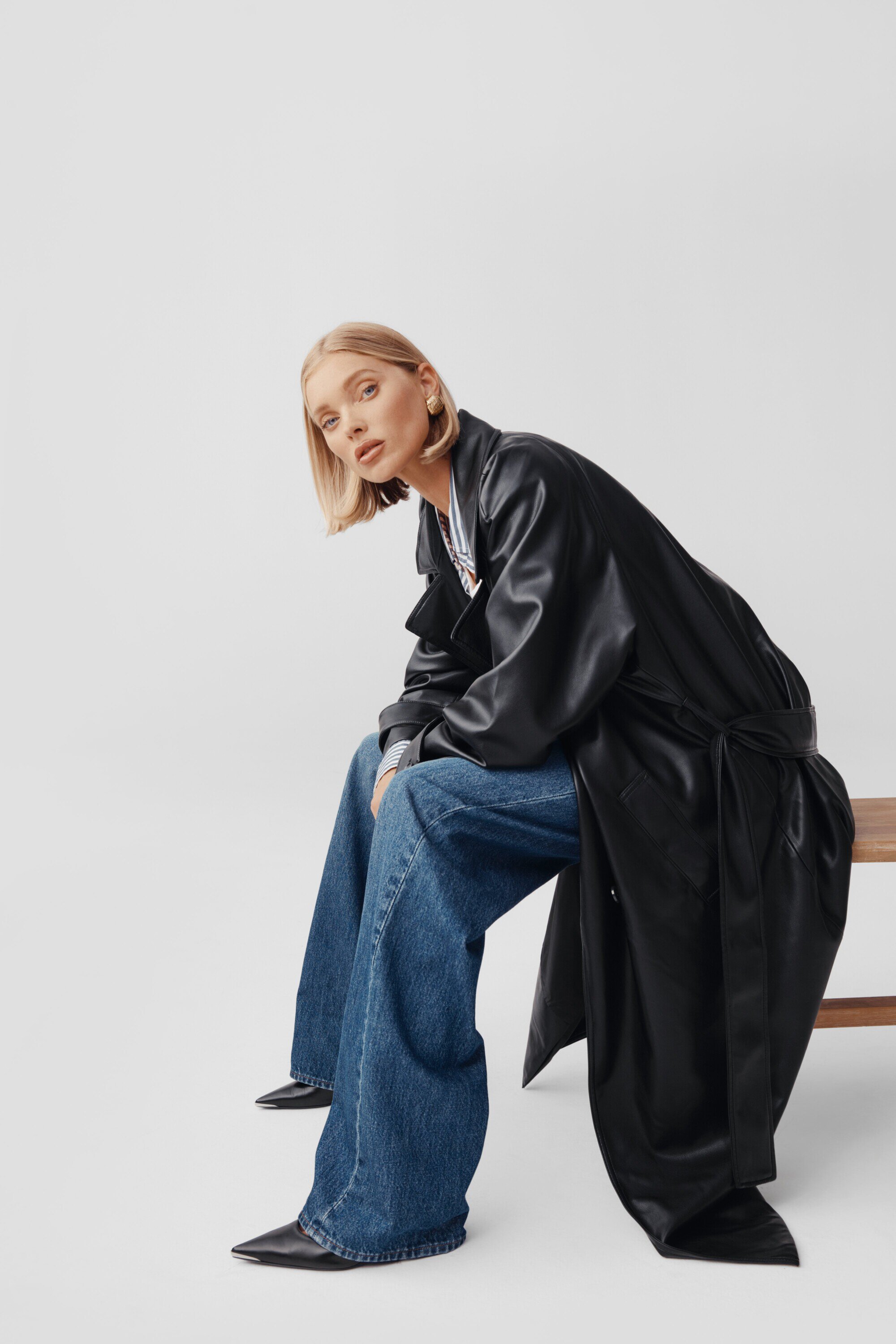 Elsa Hosk wearing a black leather trench coat and blue jeans, seated on a wooden bench against a white studio background.