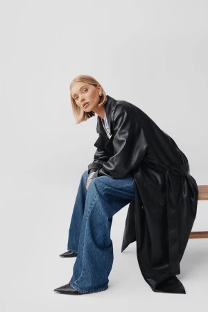 Elsa Hosk wearing a black leather trench coat and blue jeans, seated on a wooden bench against a white studio background.