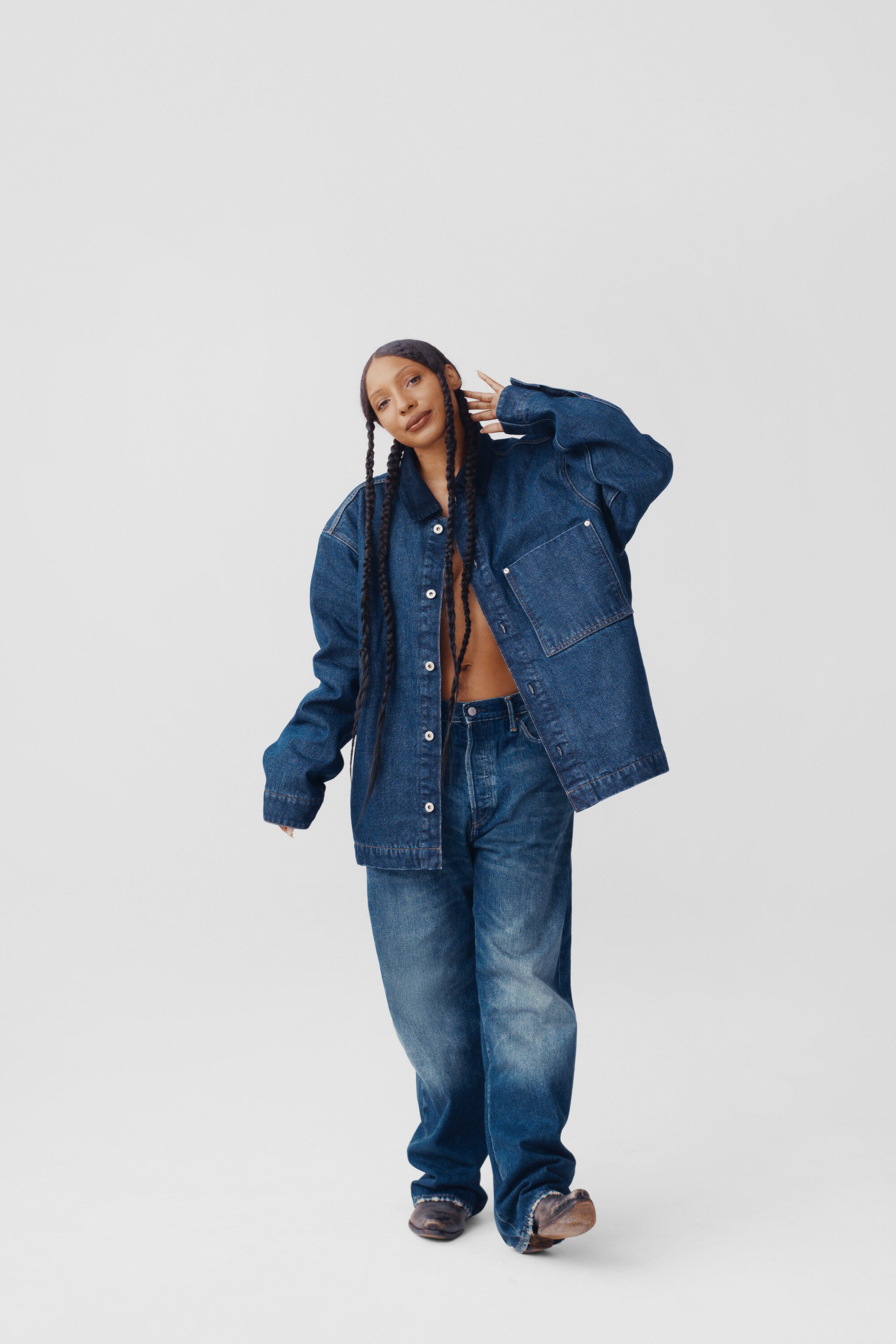 A woman poses in a denim jacket and jeans against a white background, casting a relaxed and confident vibe in a fashion forward ensemble.