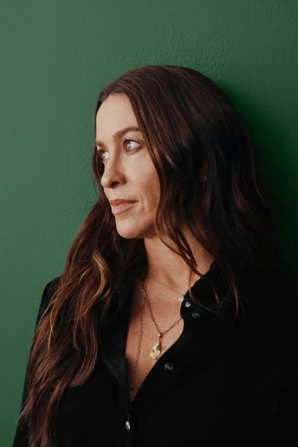 Headshot of a middle-aged woman with long brown hair, wearing a black button-down shirt and gold pendant necklace, against a green backdrop.