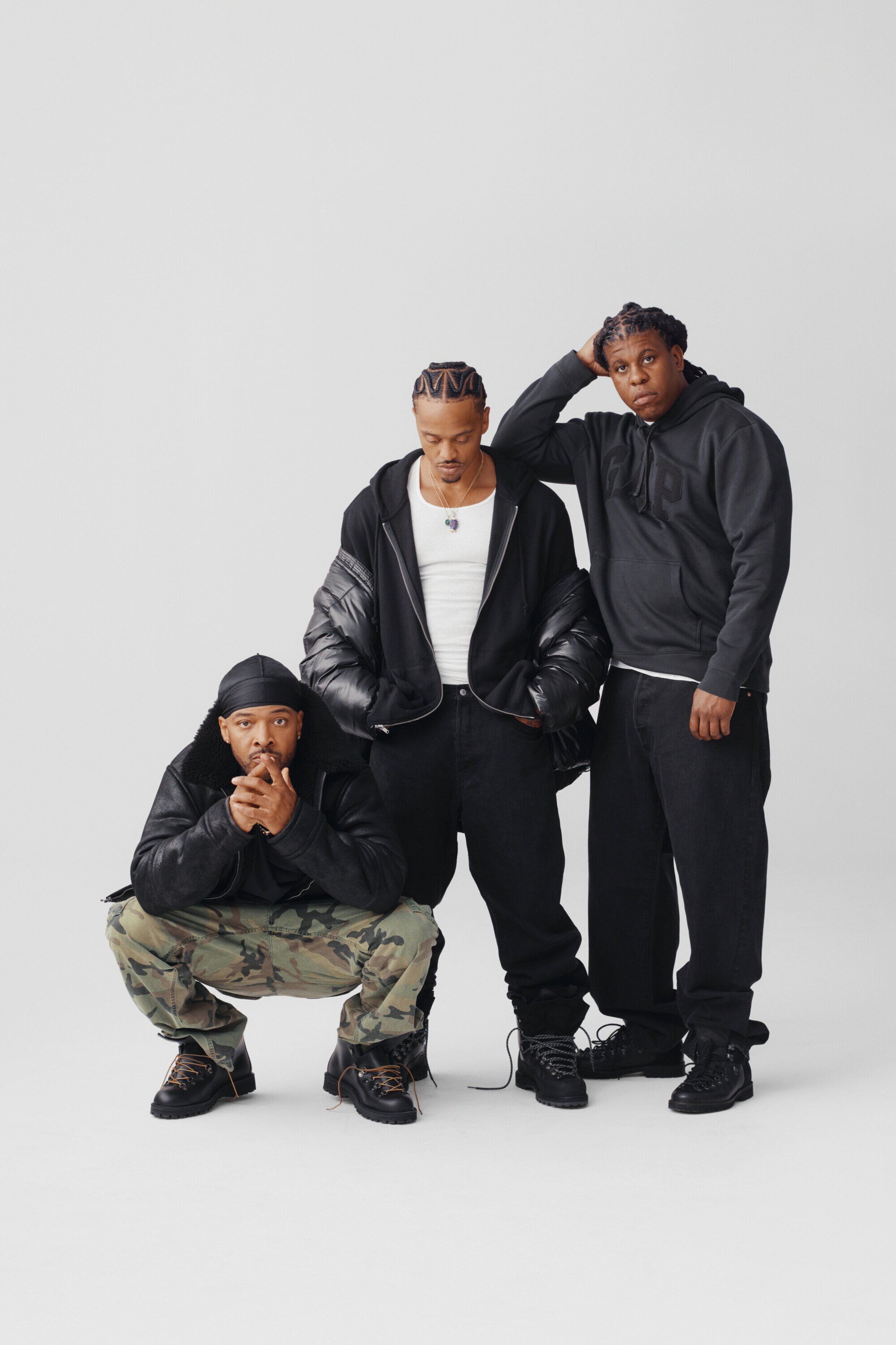 Three adult men in casual black outerwear and pants pose indoors against a neutral backdrop in a fashion-style portrait.