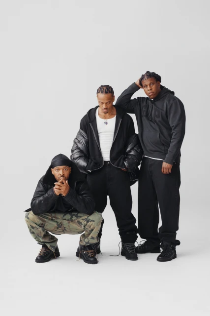 Three adult men in casual black outerwear and pants pose indoors against a neutral backdrop in a fashion-style portrait.