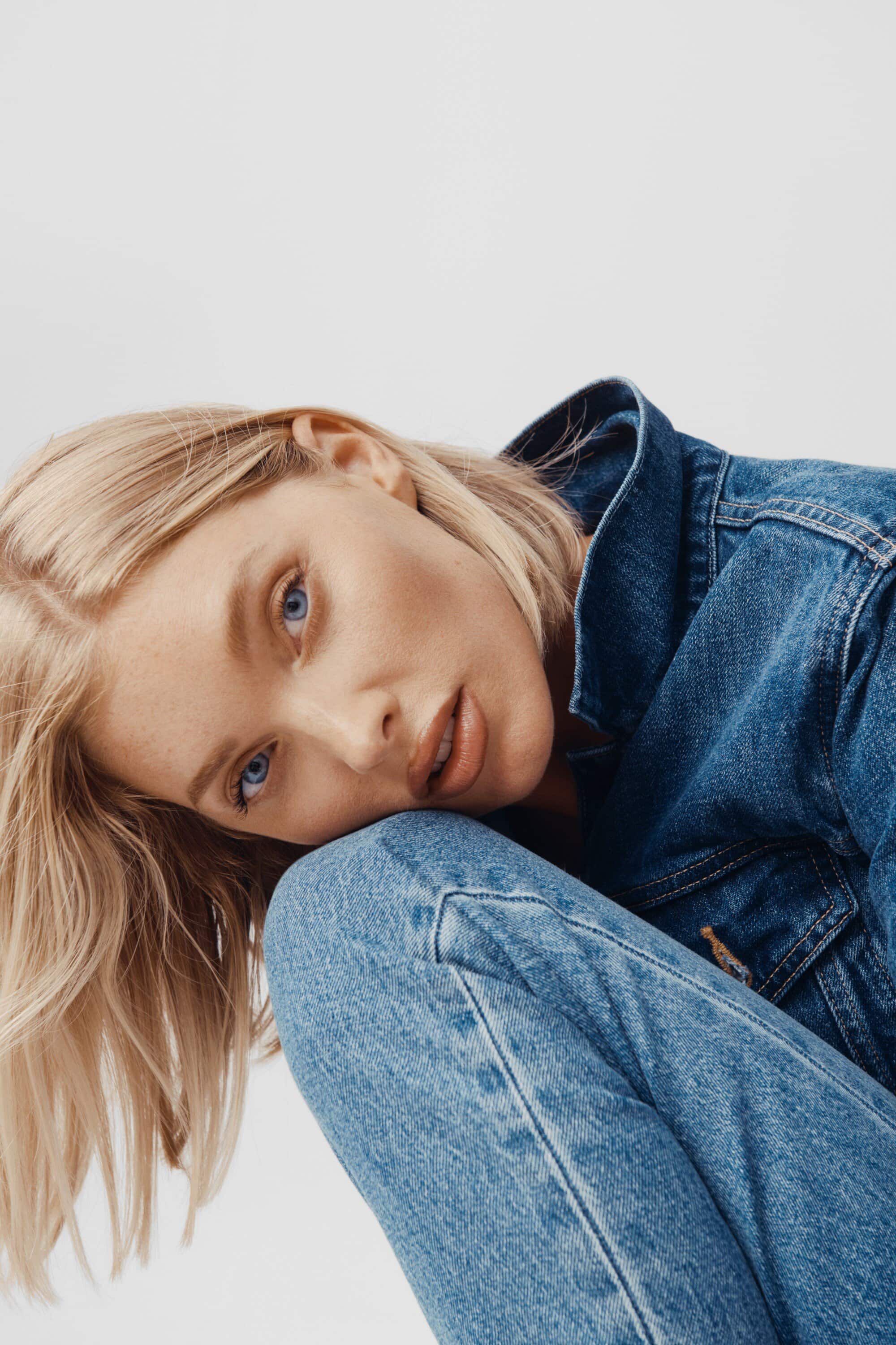 Close-up of a fair-skinned woman with blonde hair wearing a denim jacket and jeans, posing against a bright white background.