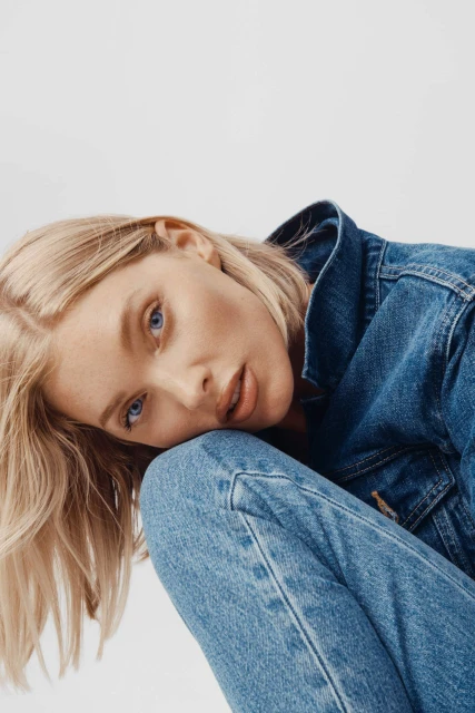 Close-up of a fair-skinned woman with blonde hair wearing a denim jacket and jeans, posing against a bright white background.